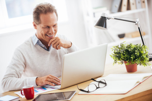 Satisfied with his work. Cheerful mature man working on laptop and smiling while sitting at his working place